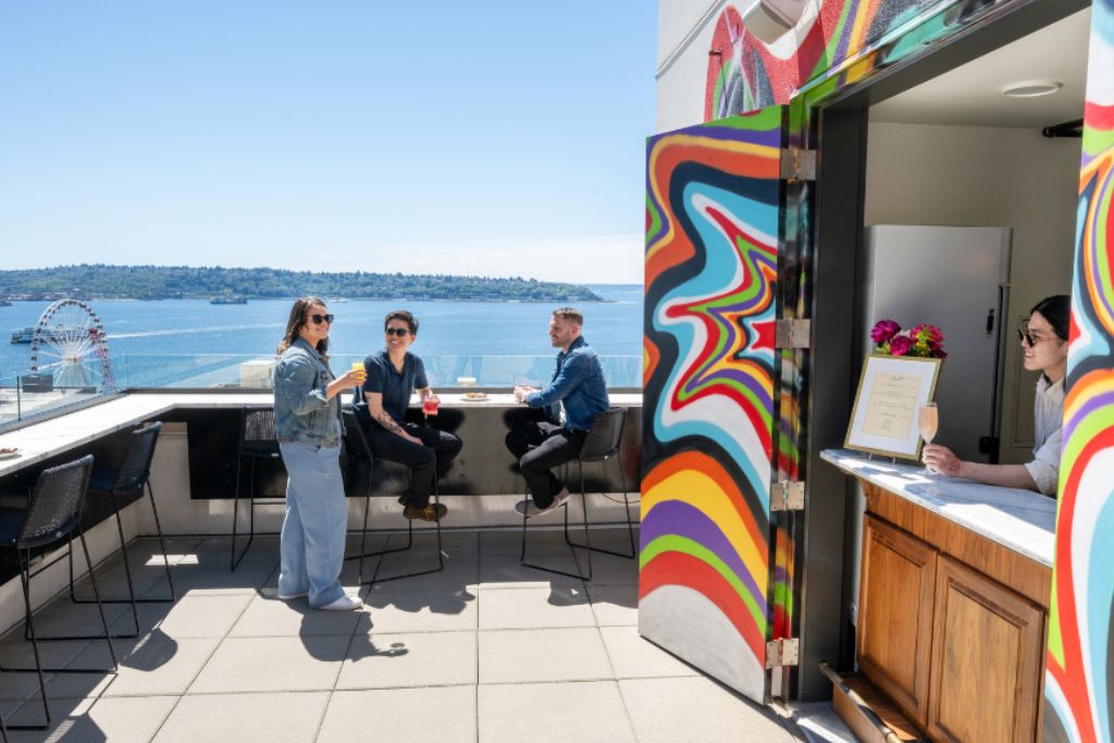 Four people enjoy drinks on a rooftop patio overlooking water and a Ferris wheel, near a vibrant, rainbow-colored kiosk where a woman stands behind a counter with a menu. The sky is clear and sunny.