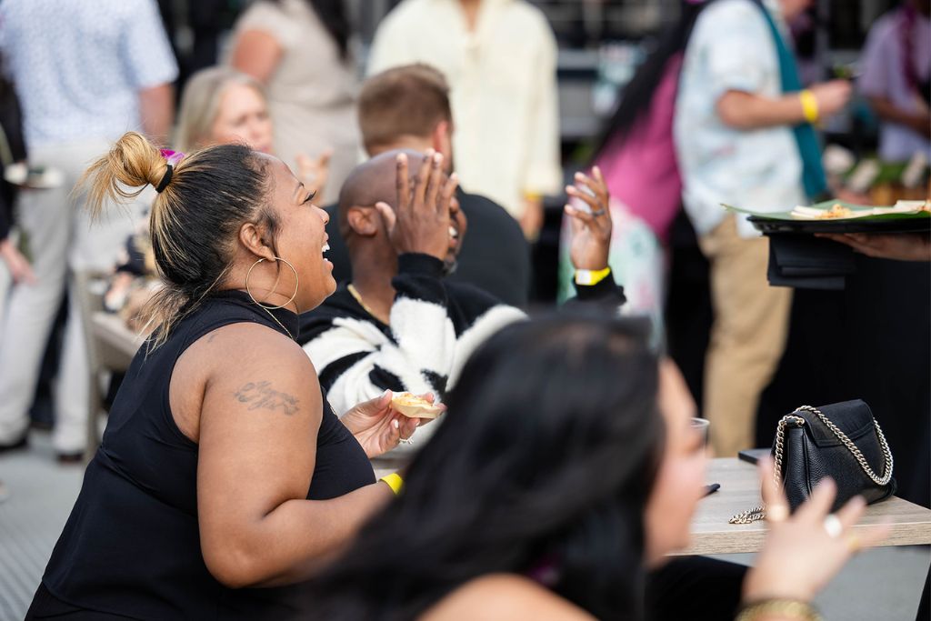 A woman with a tattoo on her arm laughs while sitting at an outdoor table with others. Nearby, a man covers his face with one hand, and people socialize in the background.
