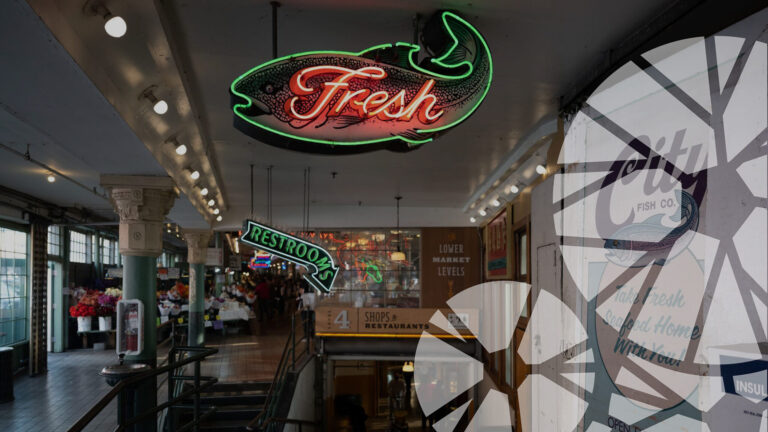 Neon fish sign reading "Fresh" hangs above stairs at an indoor market. Another neon "Restrooms" sign points left. Flower stalls and market stalls are visible in the background. A fish logo is partially reflected on the right.