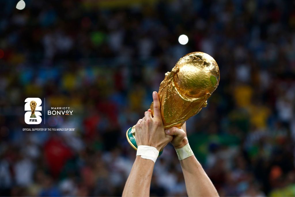 A pair of hands lifts the golden FIFA World Cup trophy in a stadium, with the crowd blurred in the background. The FIFA and Marriott Bonvoy logos appear on the left side of the image.