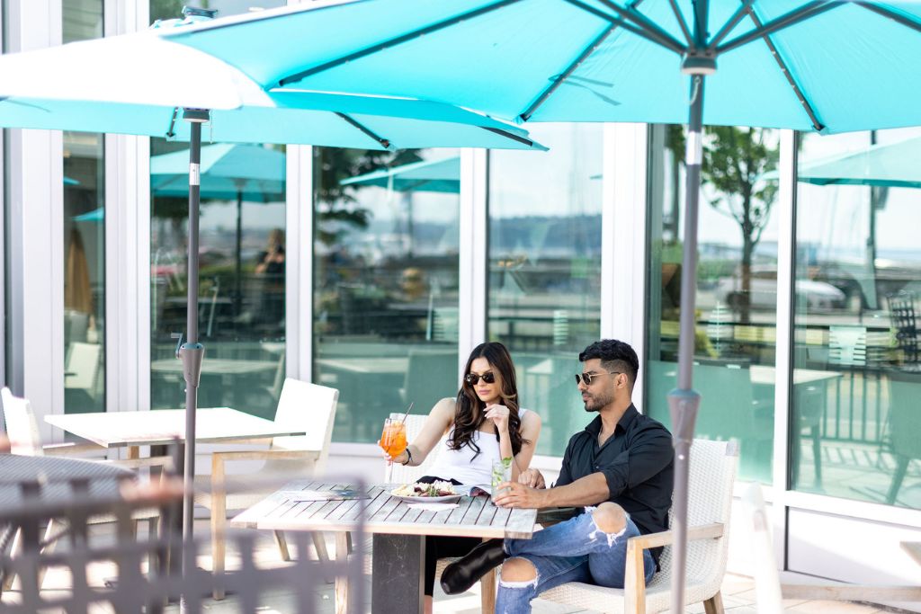 A man and woman sit at an outdoor café table under a blue umbrella, enjoying drinks and food on a sunny day. The setting is modern with glass windows and a waterfront view in the background.