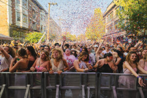 A large, energetic crowd enjoys an outdoor festival on a city street, with people dancing and smiling under a shower of colorful confetti on a sunny day. Trees and buildings line the background.