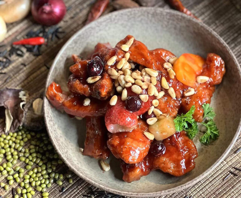 A bowl of sweet and sour pork garnished with pine nuts, cranberries, carrots, and parsley, served on a rustic mat with various spices and vegetables around.