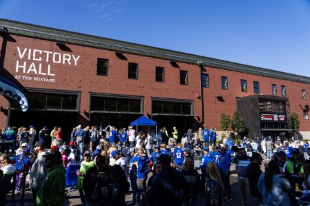 A large crowd gathers outside Victory Hall at The Boxyard on a sunny day, many wearing blue sports jerseys, indicating a lively event or sports gathering.