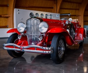 A shiny red vintage convertible car with chrome detailing is displayed indoors on a polished floor, with wooden walls and round gauges visible in the background.