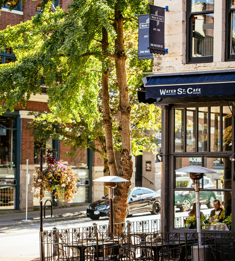 Outdoor seating area of Water St. Café on a sunny day, with empty tables and chairs, hanging flower baskets, patio heaters, and trees lining the sidewalk in a charming urban setting.