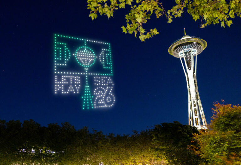 A drone light display forms a graphic and the text "FIFA World Cup 26" in the night sky near Seattle’s Space Needle, with trees framing the brightly lit tower.