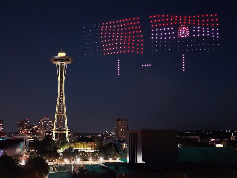 The Space Needle at night with a city skyline, while drones in the sky create a red, white, and blue American flag and the number “1” using lights.