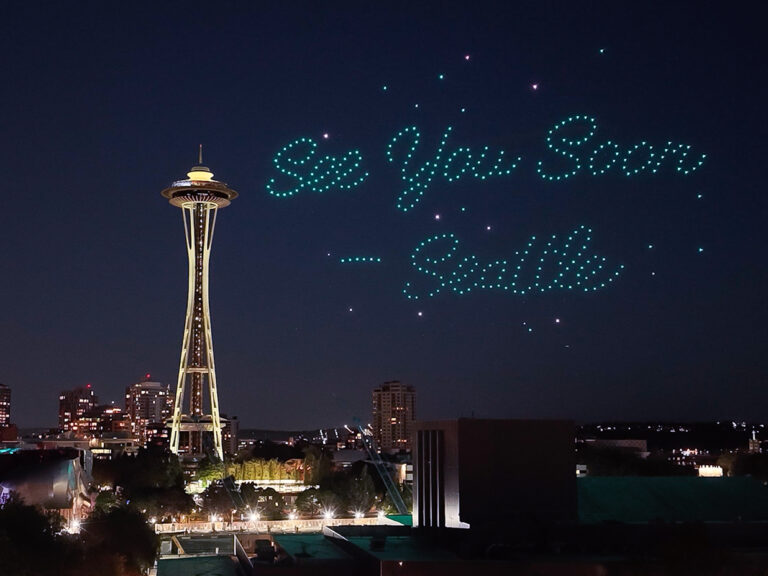 The Space Needle at night with city lights below. In the sky, blue-green drones form the words “See You Soon — Seattle.”.