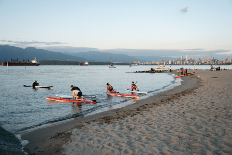 People launch paddleboards from a sandy beach at sunset. Several kayakers and paddleboarders are on the water, with a city skyline and mountains visible in the background. The sky is clear and calm.