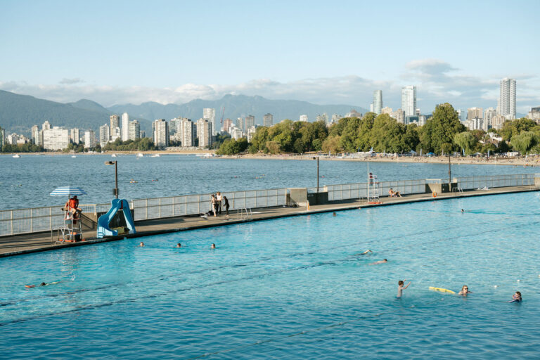 People swim in a large outdoor pool beside a waterfront boardwalk, with a city skyline and mountains in the background under a clear sky. A lifeguard sits on a stand near the pool.