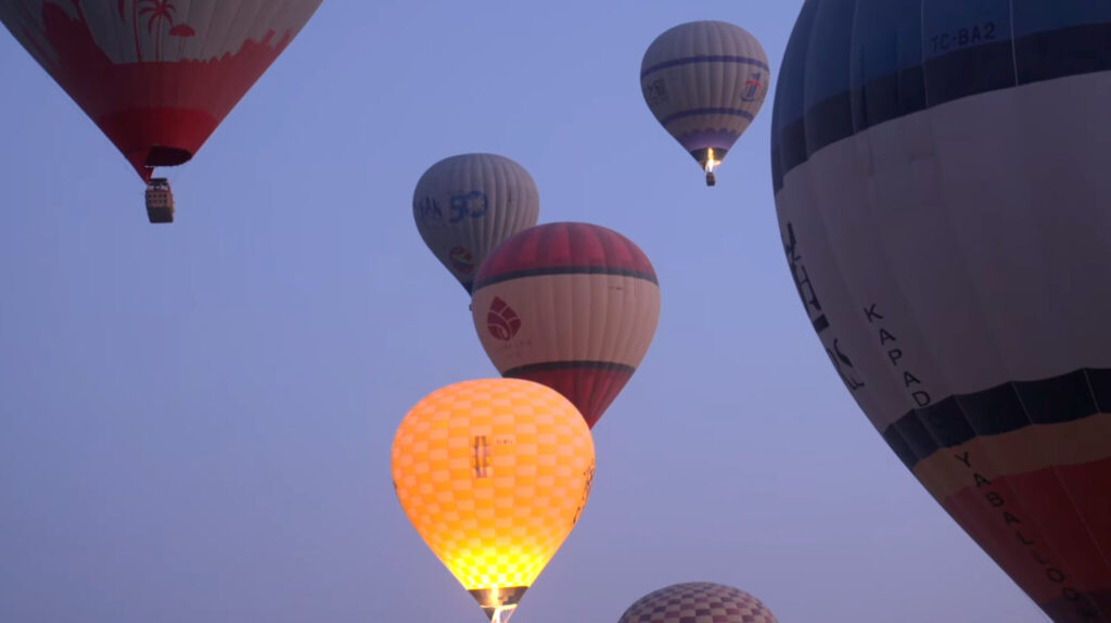 Several colorful hot air balloons float in a clear sky at dawn or dusk, with one balloon glowing brightly from its burner near the foreground.