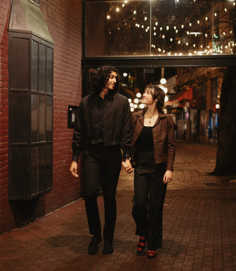 A couple holds hands and walks under string lights in a brick alleyway at night, looking at each other and smiling warmly. The scene is softly lit and cozy.