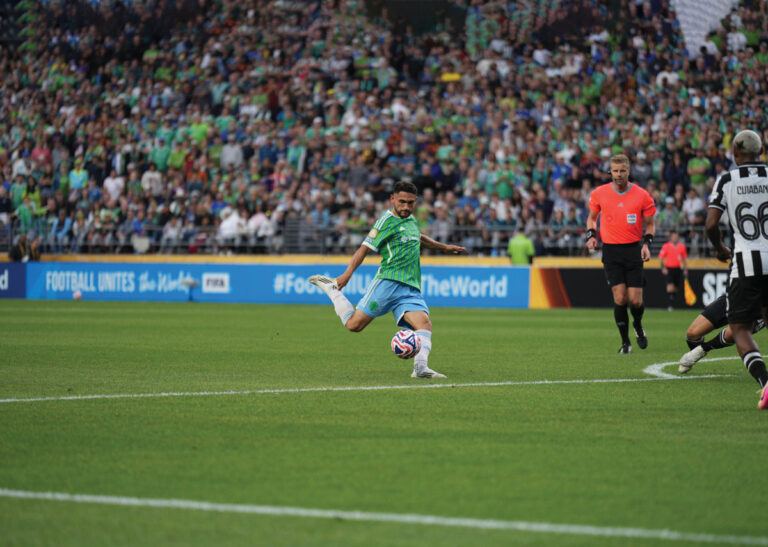 A soccer player in a green and blue uniform kicks the ball on a field during a match, with a crowd of spectators and other players in the background. An official in an orange shirt watches nearby.