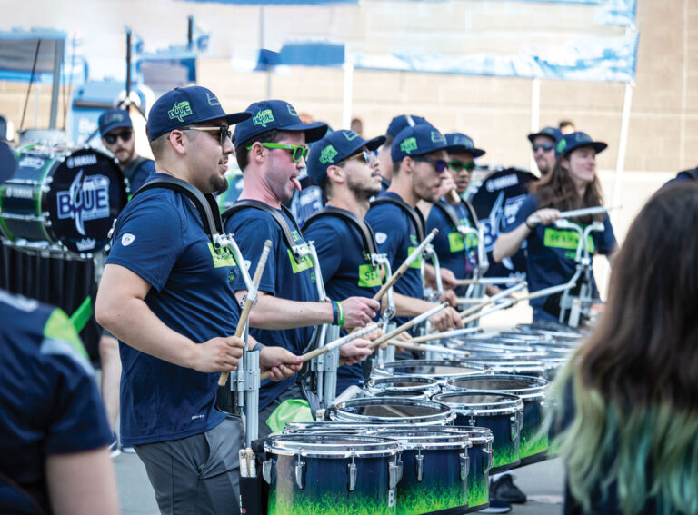 A drumline of people in matching blue shirts and hats play snare drums outdoors. Some wear sunglasses, and others watch in the background. The drums feature a blue and green gradient design.