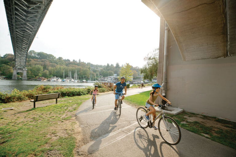 Three people wearing helmets ride bicycles on a paved path under a large bridge next to a body of water, with trees, boats, and a sunny sky in the background.