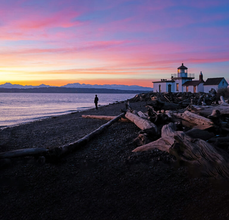 A person stands on a rocky beach with driftwood at sunset, near a lighthouse. The sky is colorful with pink, purple, and yellow hues, and distant mountains are visible across the water.