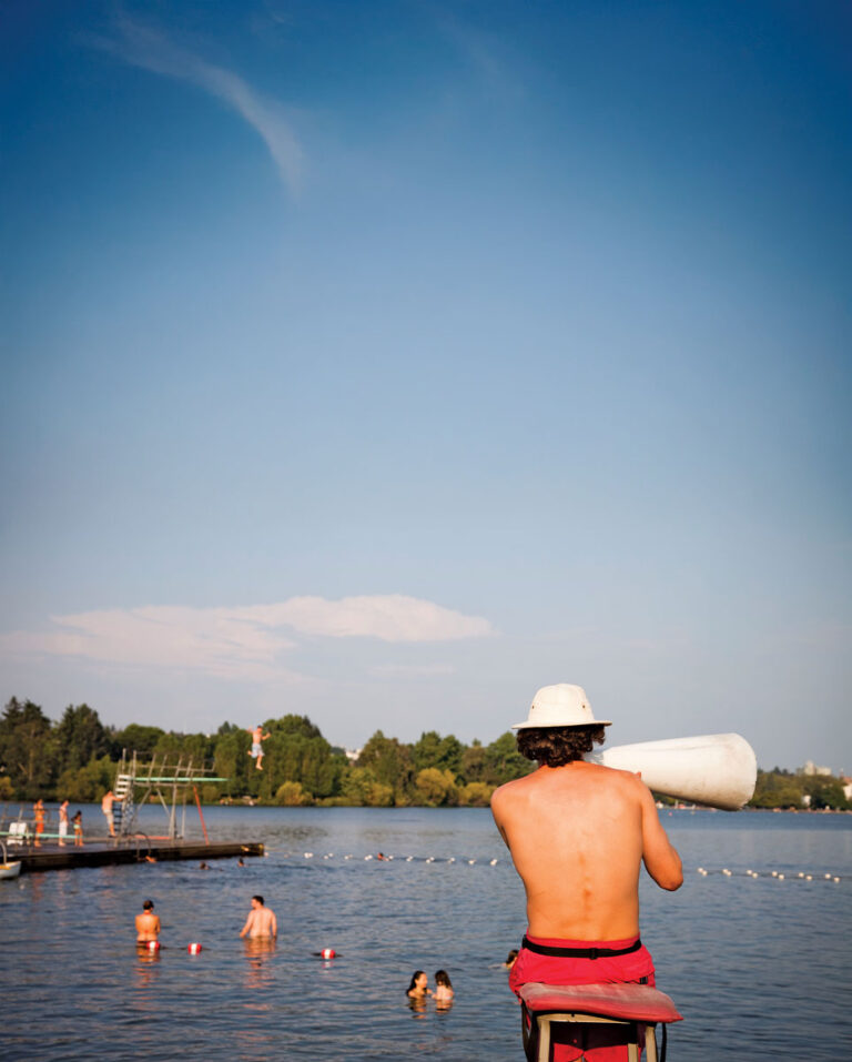 A lifeguard wearing a white hat and red shorts sits on a tall chair by a lake, holding a megaphone, watching over swimmers in the water with a dock and trees in the background under a clear blue sky.