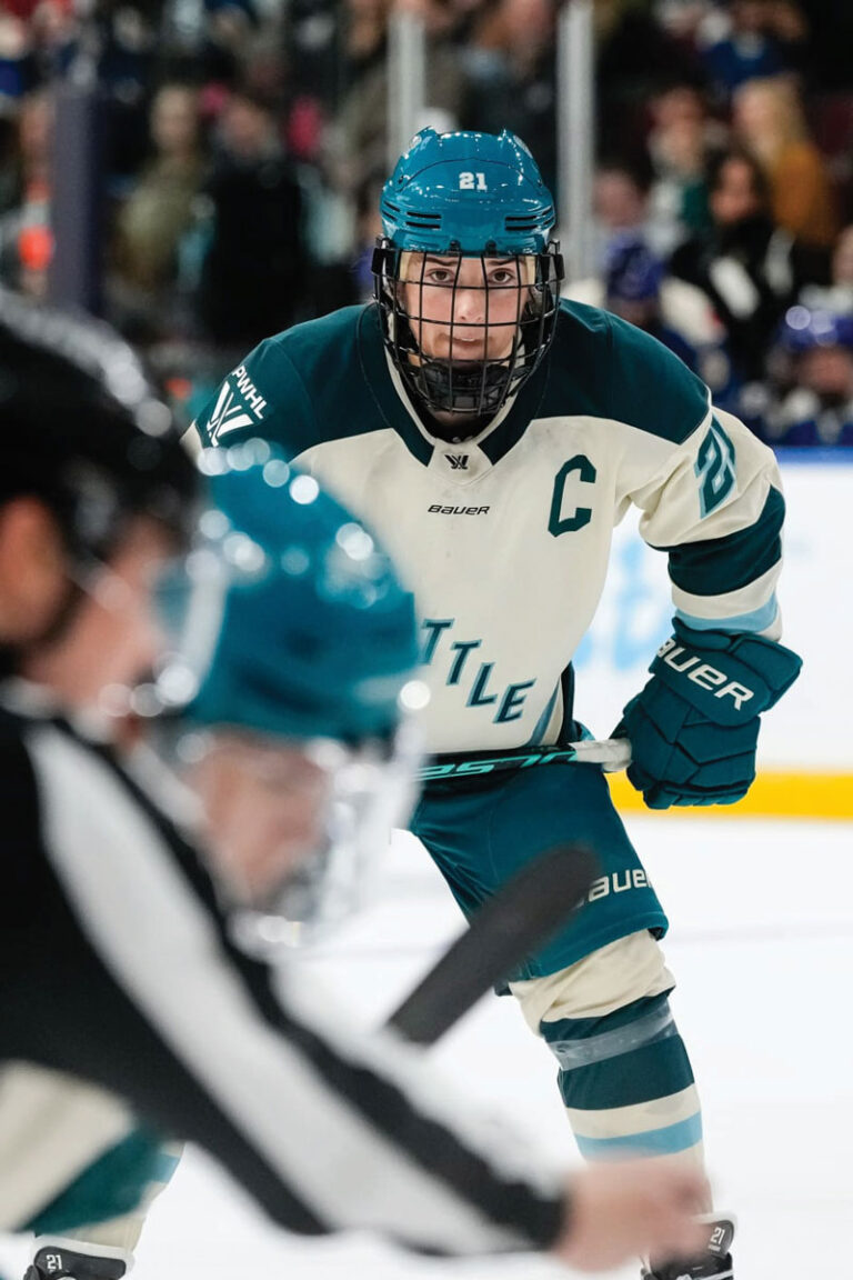 A hockey player in a white and teal uniform with a captain’s "C" on the chest focuses intently during a faceoff, with a referee and another player blurred in the foreground.