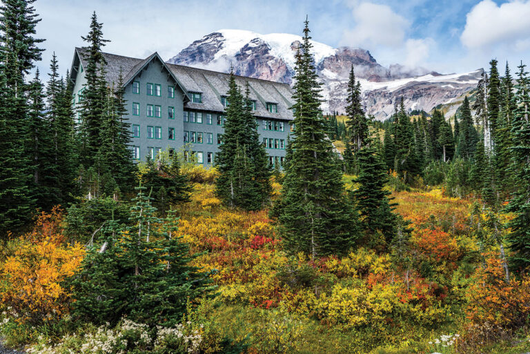 A large gray lodge with many windows stands among evergreen trees and colorful autumn foliage, with a snow-capped mountain peak rising in the background under a partly cloudy sky.