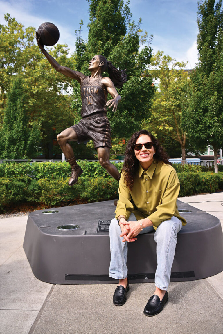A person in sunglasses and a green shirt smiles while sitting in front of a bronze basketball player statue outdoors, surrounded by greenery.