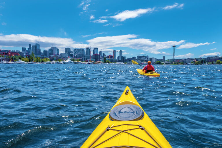 View from a yellow kayak on choppy blue water, following another kayaker toward a city skyline with skyscrapers and the Space Needle under a bright, partly cloudy sky.