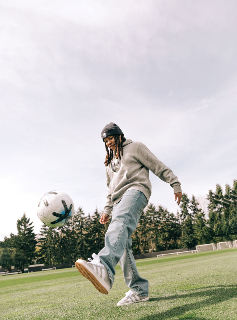 A person wearing a beanie, gray sweatshirt, and jeans kicks a soccer ball on a grassy field with trees and soccer goals in the background under a partly cloudy sky.