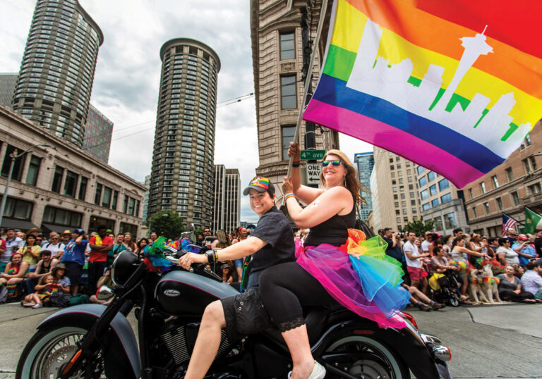 Two people in rainbow attire ride a motorcycle in a city parade, one holding a rainbow flag with a white silhouette of Seattle’s Space Needle. A large crowd watches from the sidewalk, with tall buildings in the background.