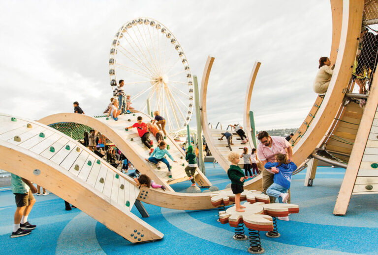 Children and adults play on modern wooden playground structures with blue flooring. A large Ferris wheel rises in the background under a cloudy sky, and the area is busy with people enjoying the park.