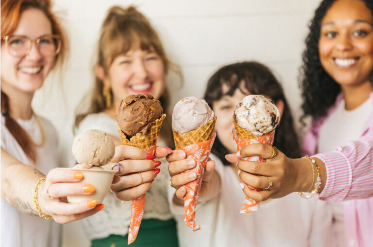 Four smiling people hold out various ice cream cones and a cup toward the camera, displaying different flavors in waffle cones with patterned orange wrappers. The focus is on the ice cream, with faces softly blurred in the background.