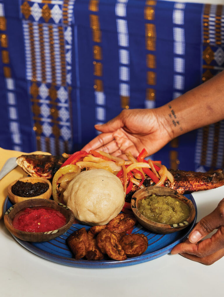 A hand presents a blue plate with a whole grilled fish, a round serving of fufu, fried plantains, and three small bowls of red, dark, and green sauces, set against a blue patterned cloth background.