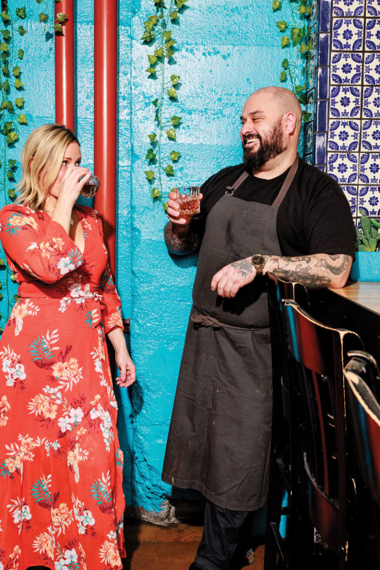 A woman in a floral red dress and a man in a black apron stand by a blue wall, smiling and drinking together, with decorative tiles and hanging plants in the background.