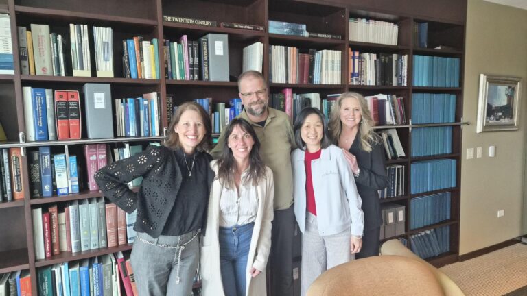 Five people stand smiling in front of a large bookshelf filled with books in an office or library setting. Four women and one man are casually dressed, appearing happy and relaxed together.