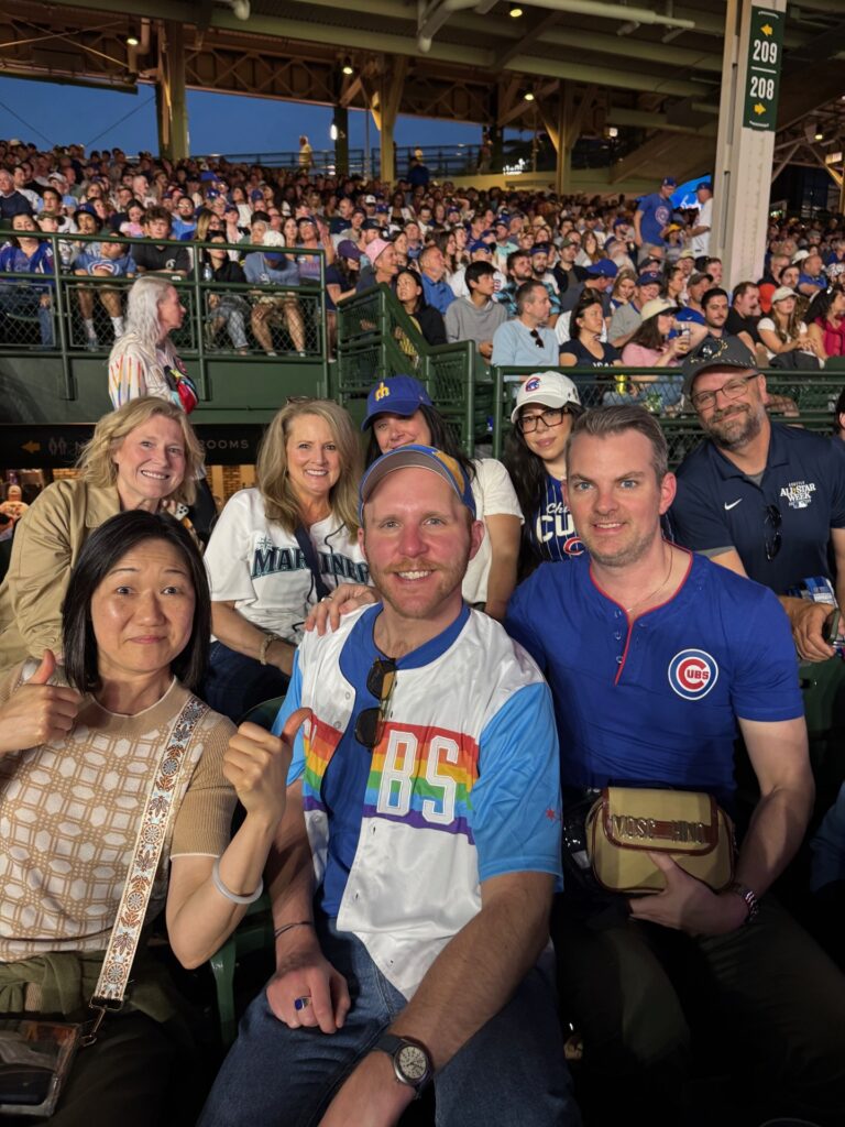 A group of smiling people sit in stadium seats at a baseball game, wearing Cubs gear. The stands behind them are packed with fans under bright stadium lights.