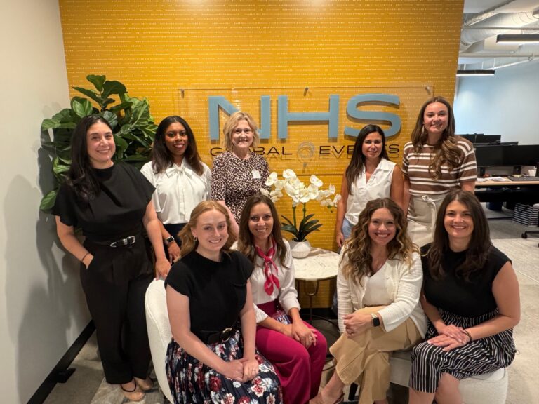 Nine women, some seated and some standing, pose and smile in an office in front of a yellow wall with "NHS GLOBAL EVENTS" text, beside a white orchid and green plant.
