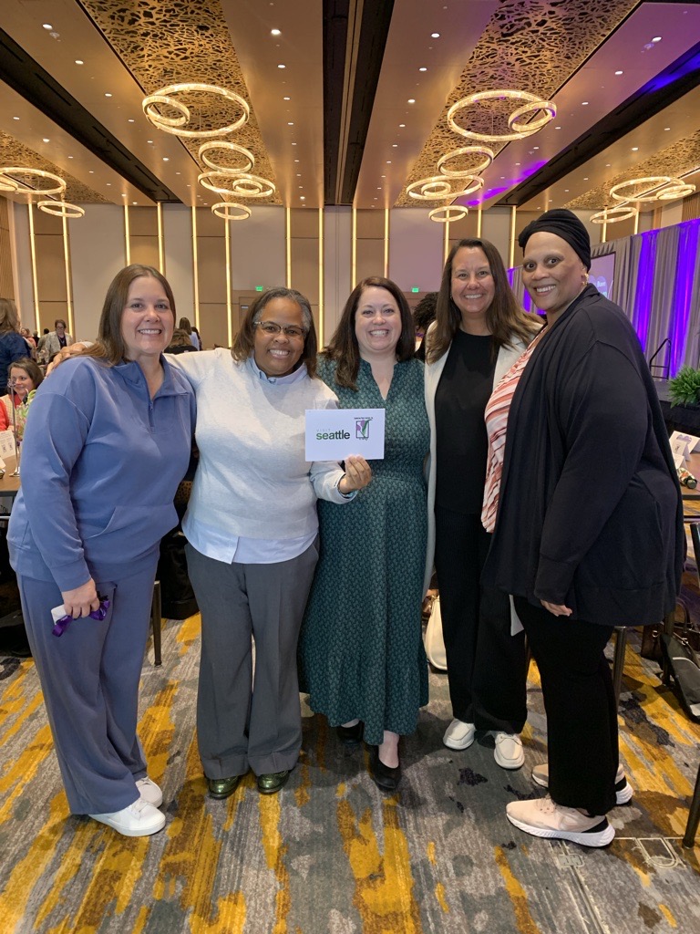 Five women stand together and smile at an indoor event, holding a “Seattle” sign. The room has golden lighting, round tables, and decorative ceiling lights.