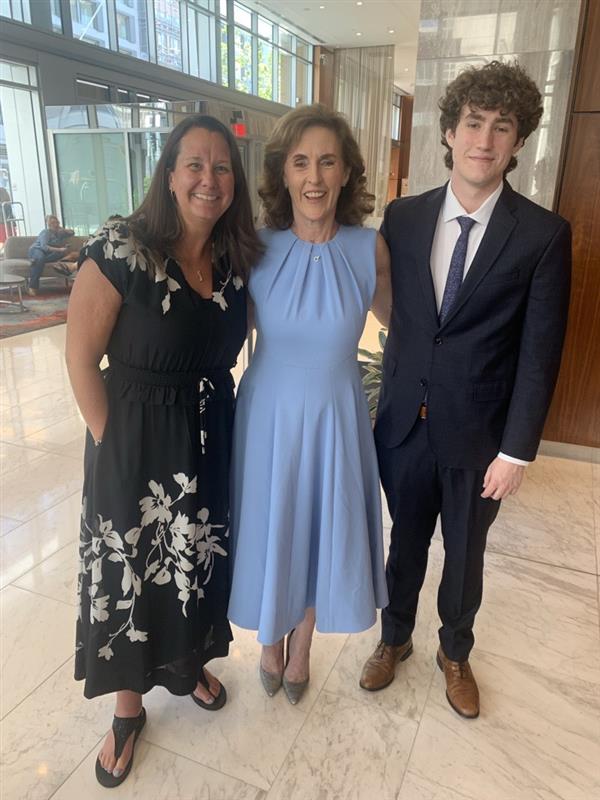 Three people pose together indoors: a woman in a black floral dress, a woman in a light blue dress, and a young man in a suit. They are all smiling and standing on a marble floor in a bright, modern lobby.