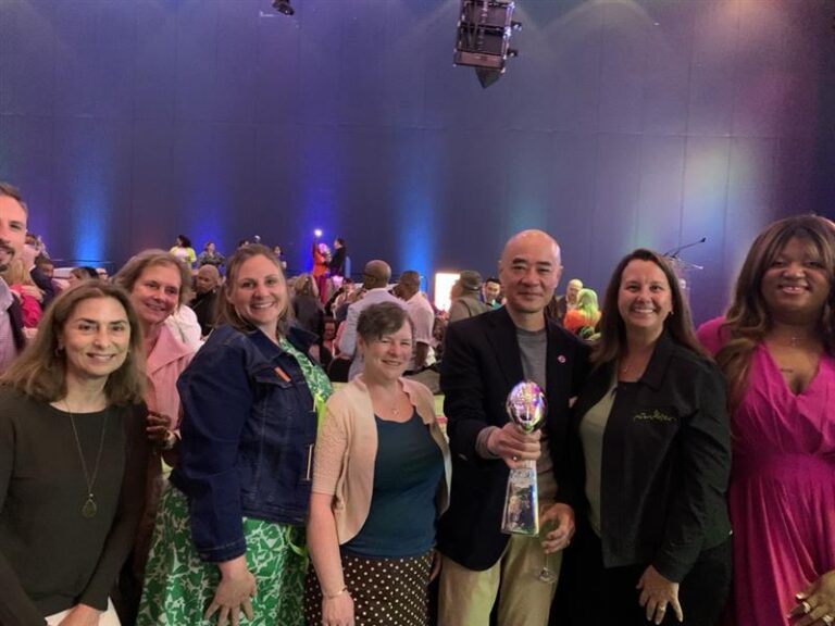 A group of people pose together at an indoor event. One person in the center holds a glass trophy. Colorful lights illuminate the background, and everyone is smiling at the camera.