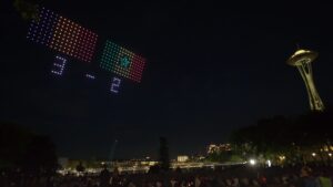 Colorful drones form the score "3 - 2" and the flags of France and Senegal in the night sky above a city crowd, with the Space Needle visible in the background—a dazzling Visit Seattle moment brought to life by a spectacular FIFA World Cup drone scoreboard.