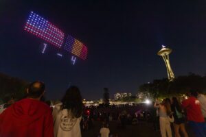 People watch a nighttime drone light show near the Space Needle, where drones form a scoreboard and display the numbers "1 - 4" in the sky—a spectacular moment celebrating Seattle’s role as a FIFA World Cup city. Visit Seattle to experience unforgettable sights like this innovative drone scoreboard lighting up the night.