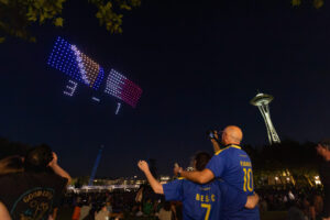 A group watches a night sky Visit Seattle FIFA World Cup drone scoreboard show, displaying a blue and red "3 - 1" near the Space Needle in Seattle. Some people wear blue jerseys and take photos.