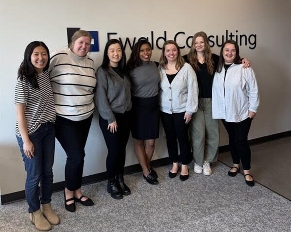 Seven women stand smiling in front of a wall with the Ewald Consulting logo. They are dressed in business casual attire and standing closely together in an office setting.