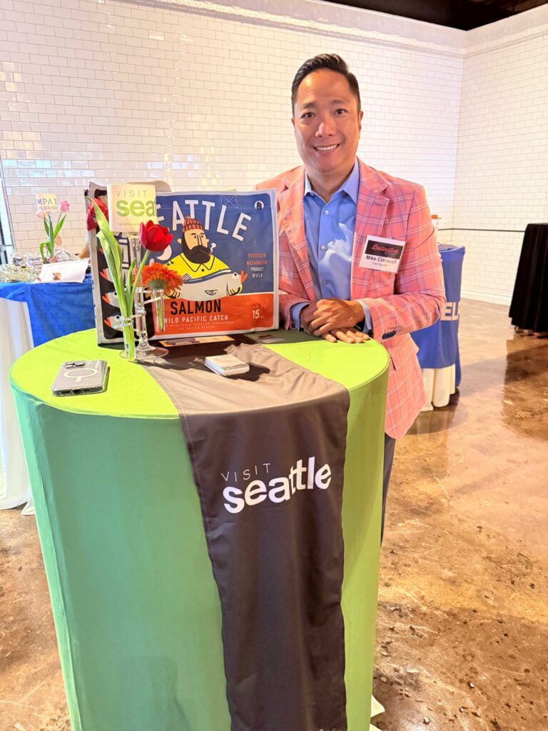 A man in a pink blazer stands behind a green table with a "Visit Seattle" cloth, a salmon sign, flowers, and brochures. He smiles at the camera in a bright indoor setting with white tile walls.