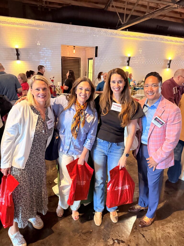Four people stand indoors at an event, smiling at the camera, each holding a red gift bag. The background shows other attendees and tables in a well-lit room with white brick walls.