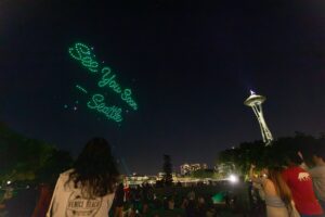 A night sky in Seattle with green drones forming the words "See you soon - Emily" near the Space Needle, as people watch from a grassy park.