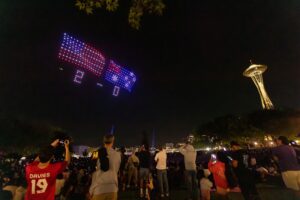 A crowd watches drones form a US flag and score “2-0” in the night sky near the Space Needle. People are taking photos and videos of the light show.