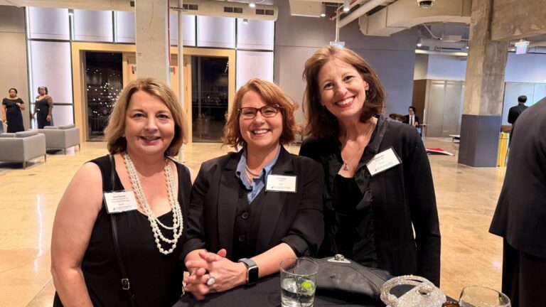 Three women in business attire smile at a formal indoor event, standing together near a cocktail table with drinks. Name tags are visible on their jackets. The background features modern decor and other attendees.