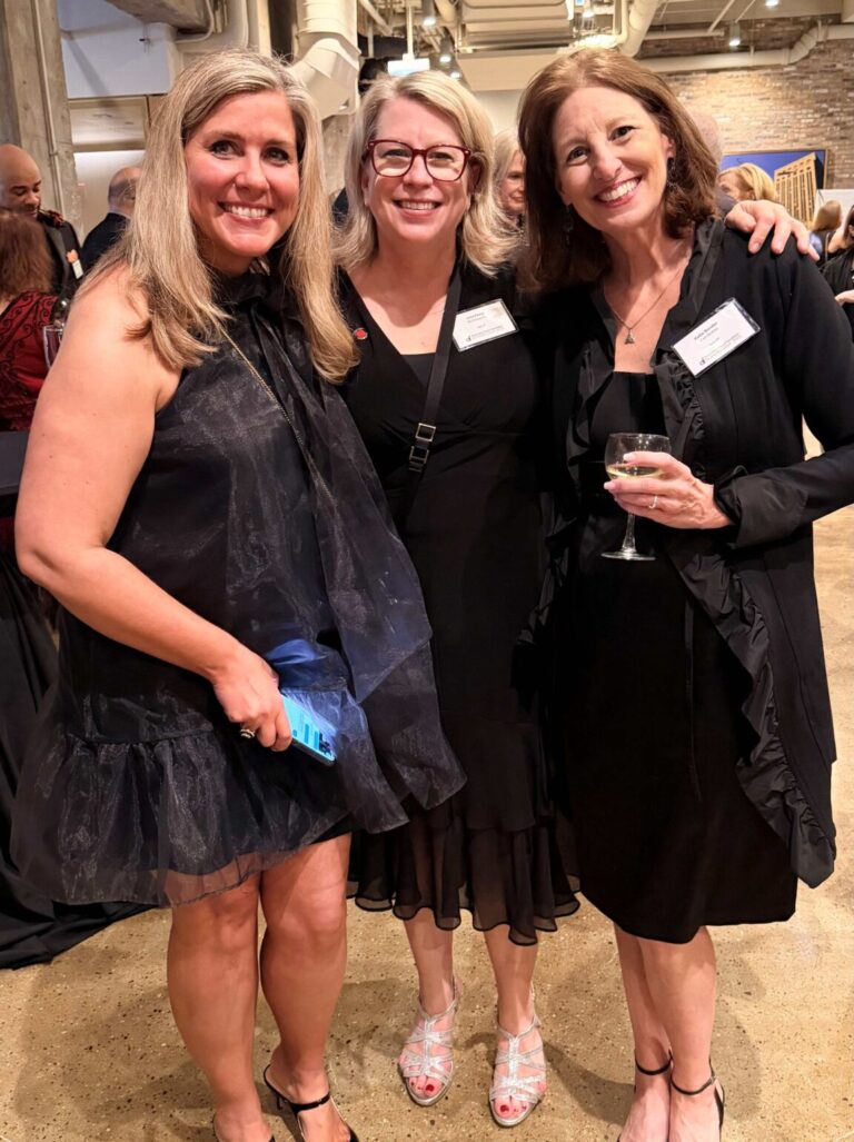 Three women dressed in formal attire smile and pose together at an indoor event. One holds a drink, another holds a phone. Other guests and exposed brick walls are visible in the background.