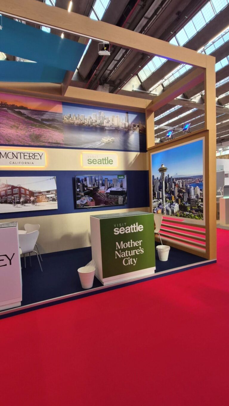 A trade show booth with large photos of Seattle and Monterey, a green sign reading "Visit Seattle Mother Nature's City," white chairs, counters, and red carpet flooring under a high ceiling with skylights.