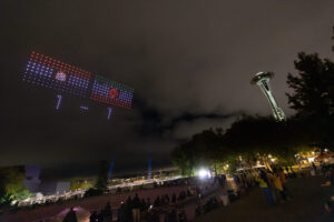 Drones form two rectangular flag patterns in the night sky near Seattle’s Space Needle, as people watch from below during a Visit Seattle FIFA World Cup drone scoreboard event.